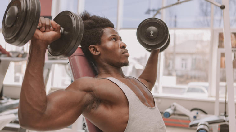 Man sitting in gym pressing dumbbells overhead